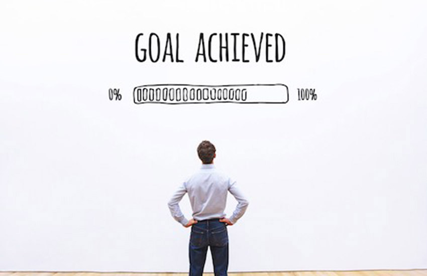 man with his hands on his hips, looking at a wall that says, "Goal Achieved," written above a progress bar.